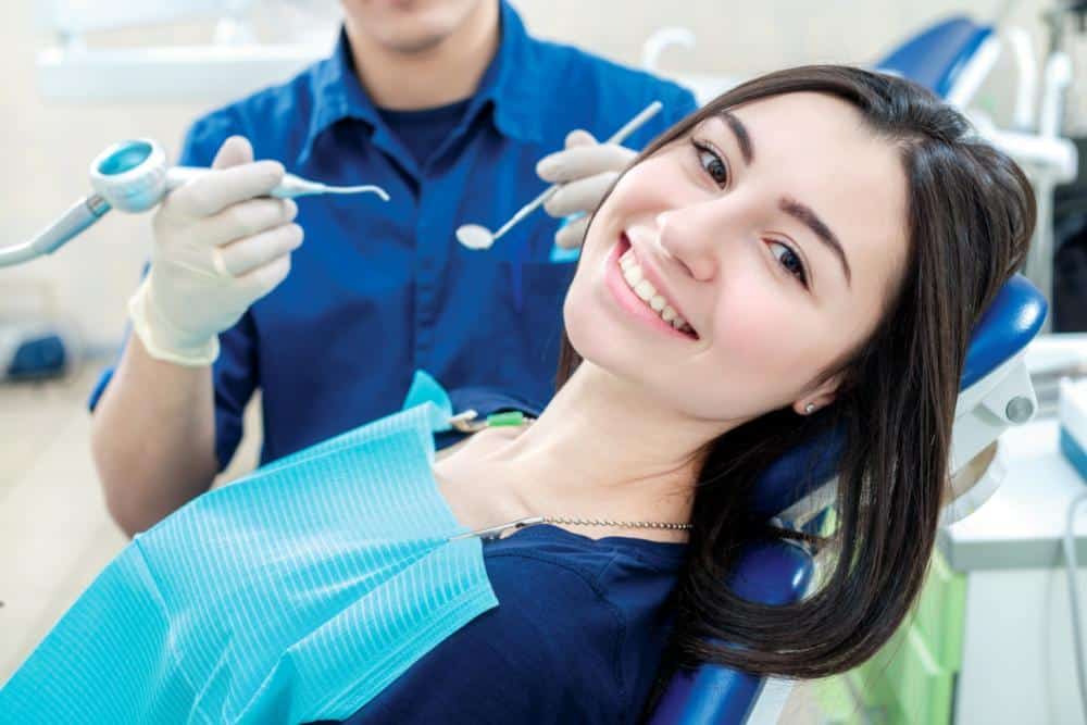Smiling female patient in dental chair receiving examination from dentist in blue uniform with tools.