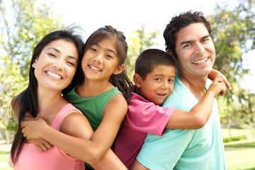 Happy family of four smiling outdoors in park setting, embracing together.