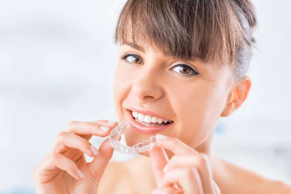 Young woman smiling while holding clear dental aligners against white background.