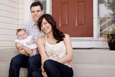 Family of three sitting on front porch steps of brick home, smiling at camera.