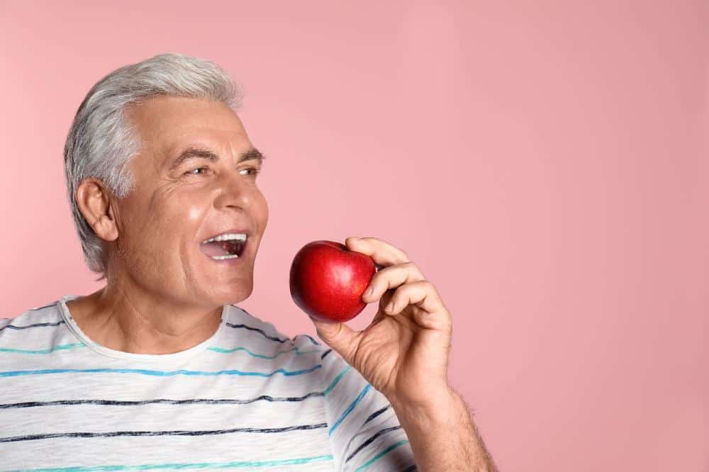 Senior man holding red apple against pink background, smiling.