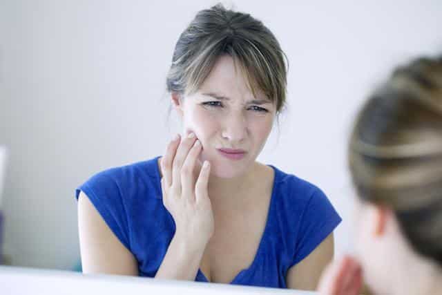 Woman in blue shirt touching cheek in pain, dental office setting with mirror visible.