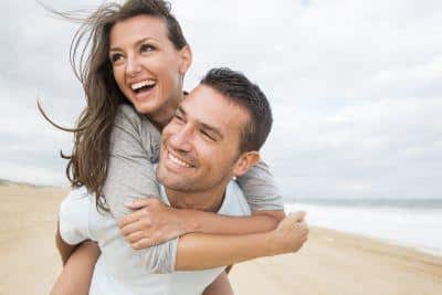 Happy couple enjoying beach time together, man giving woman piggyback ride by ocean.