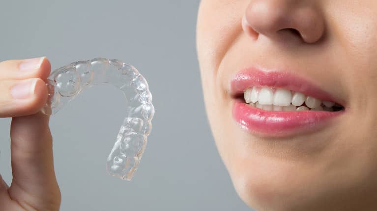 Person holding clear dental aligner near smiling teeth against gray background.