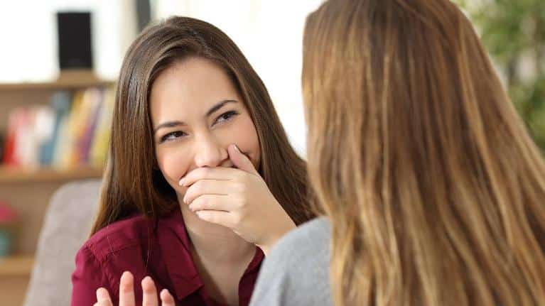 Two women having conversation indoors, one covering mouth while smiling, dental office setting with shelves background.