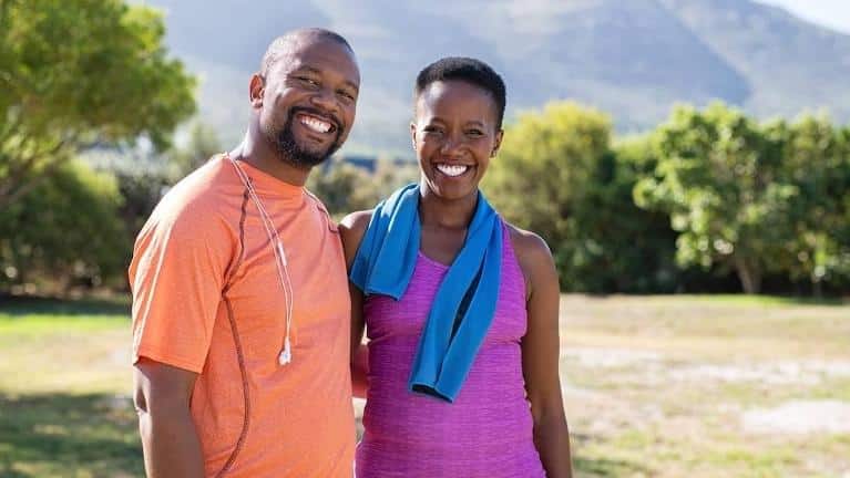 Two smiling people standing together outdoors in a park setting with mountains in background.