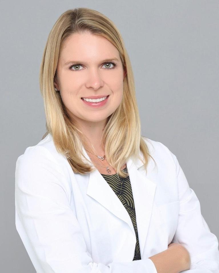 Professional headshot of female dentist in white coat against gray background.