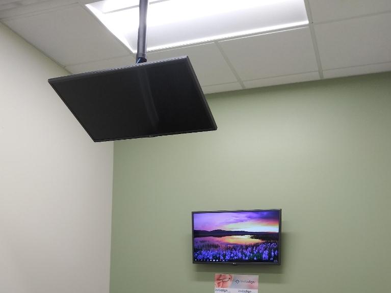 Dental office waiting room with mounted TV displaying scenic landscape on light green wall below black ceiling-mounted speaker.