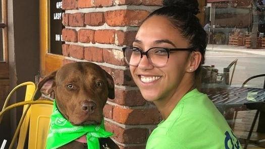 Young woman wearing glasses and green shirt smiling while holding brown dog with green bow indoors.