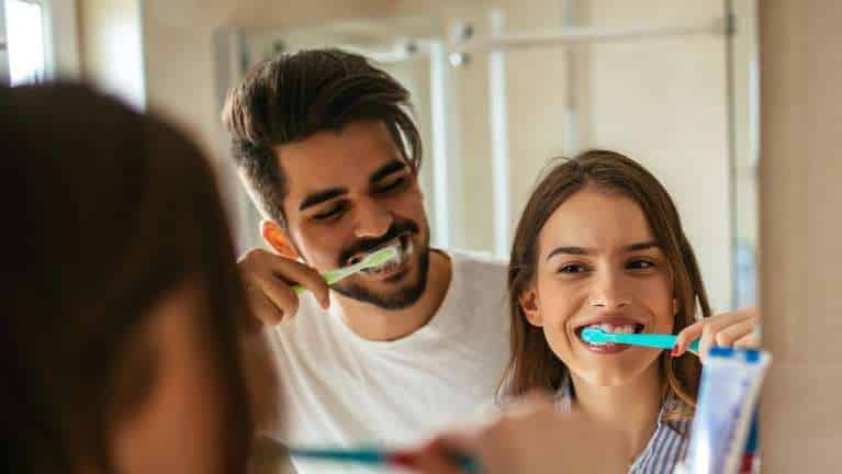 Couple brushing teeth together at bathroom mirror, smiling and practicing daily oral hygiene routine.