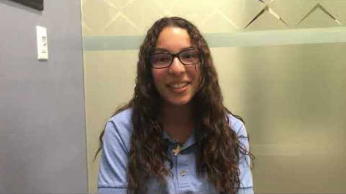 Woman wearing glasses and blue shirt smiling at camera in dental office setting.